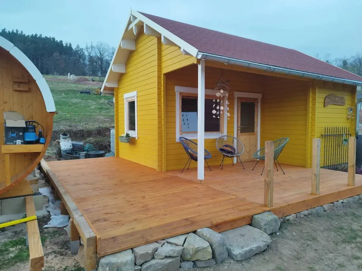 Ein gelber Holzschuppen mit Terrasse, ideal zum schuppen selber bauen. Daneben eine Holzfasssauna.