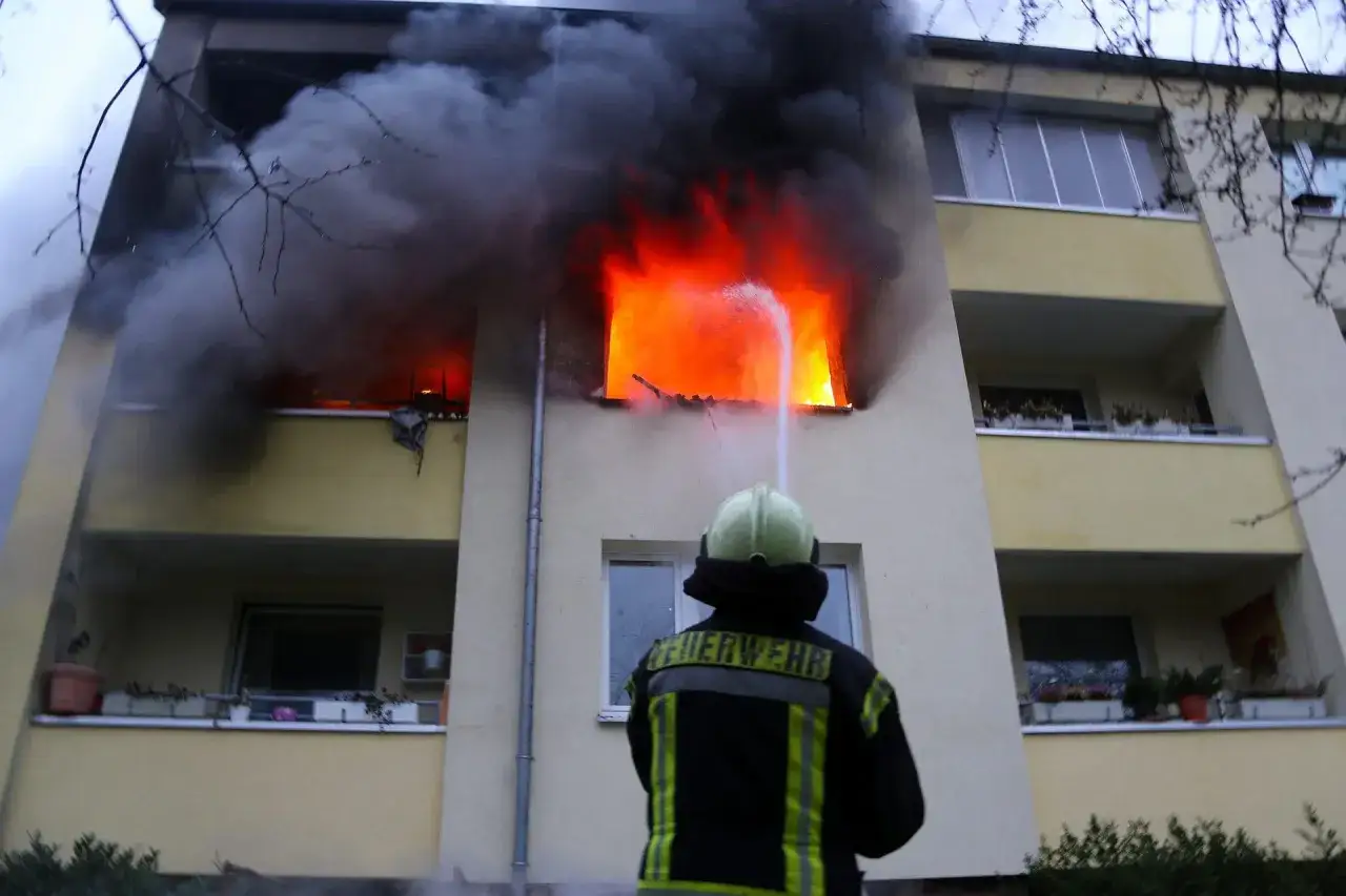 Feuerwehr l&ouml;scht Brand in Wohnung. Dunkler Rauch und helle Flammen lodern aus einem Fenster. Ein Feuerwehrmann mit Helm und Jacke k&auml;mpft gegen die Hitze.