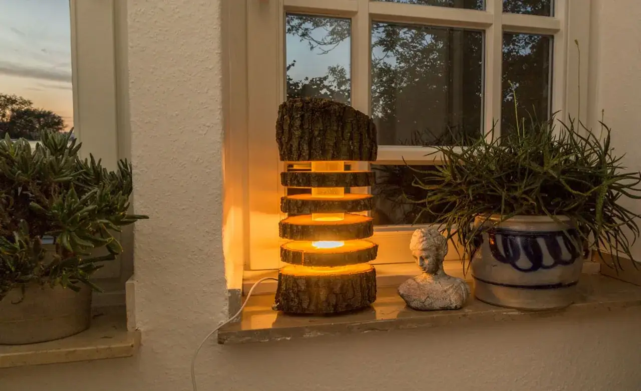 Eine rustikale Lampe, die man aus einem Baumstamm selber machen kann, leuchtet warm auf einem Fensterbrett neben Pflanzen.