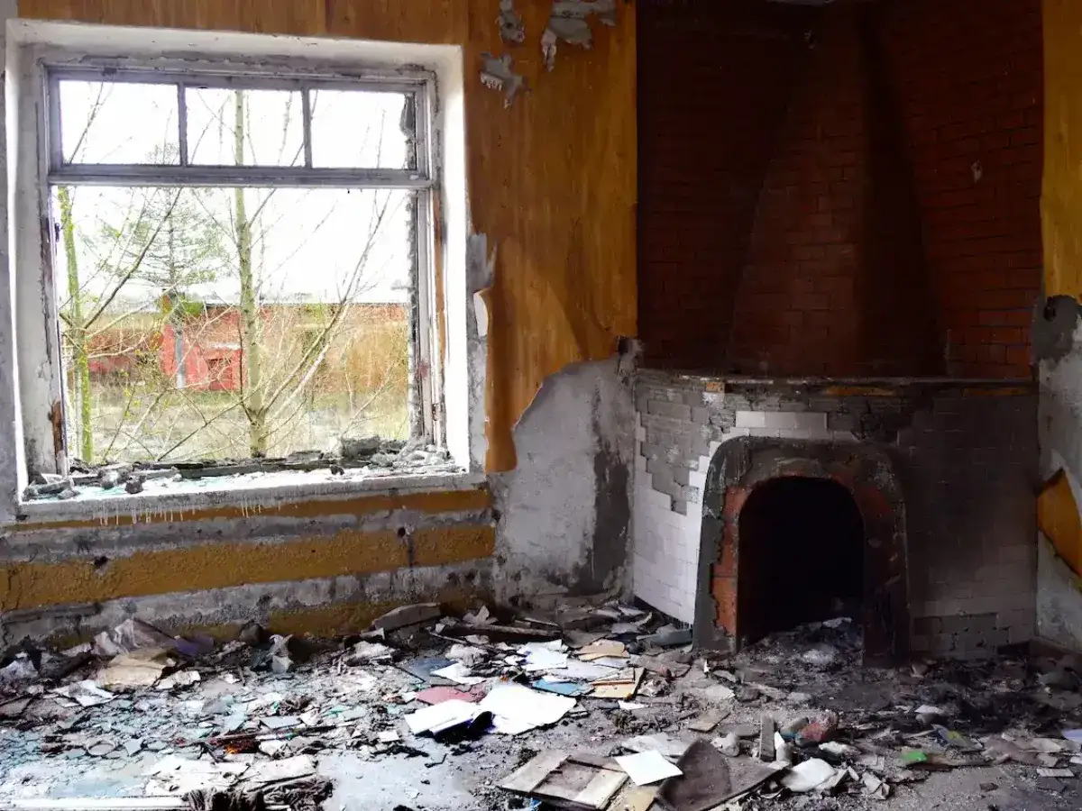 Verlassenes Zimmer mit zerbrochenem Fenster und Kamin. Der Boden ist mit Schutt bedeckt. Man kann fast den geruch nach brand giftig riechen.