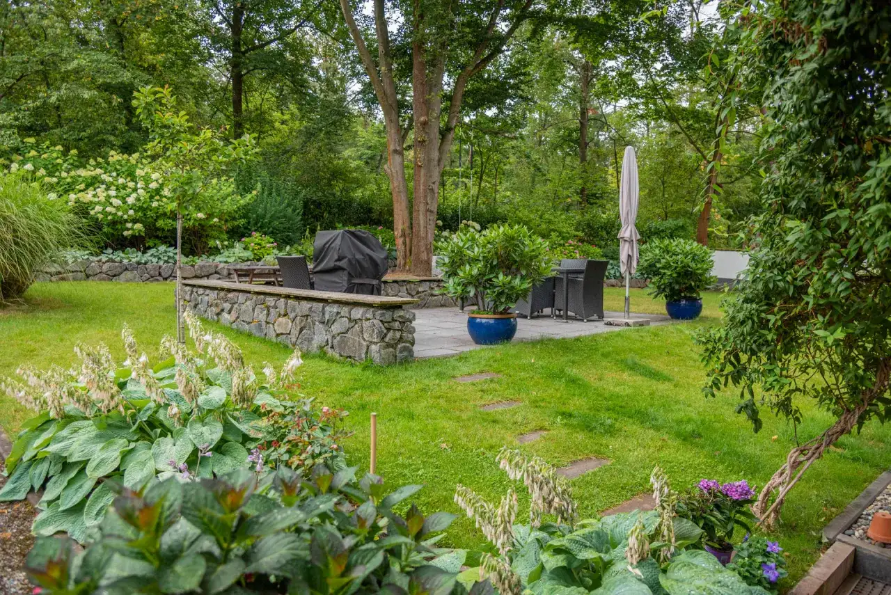 Moderner Garten am Hang mit Steinterrasse, Grill, Sitzgruppe und &uuml;ppiger Bepflanzung.