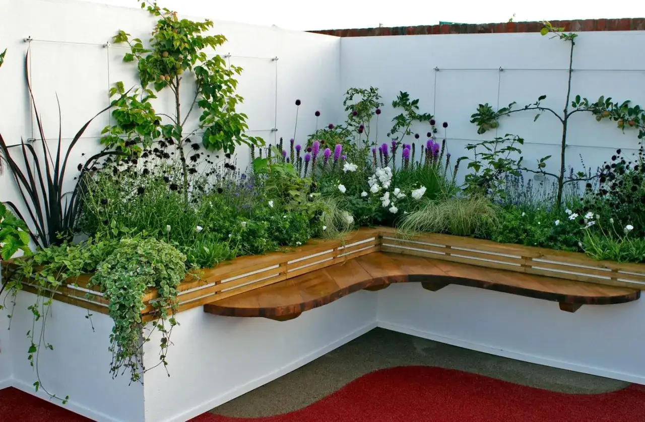 Ein moderner, kleiner Garten mit einer geschwungenen Holzbank, umgeben von &uuml;ppiger Bepflanzung mit lila und wei&szlig;en Bl&uuml;ten sowie Gr&auml;sern.