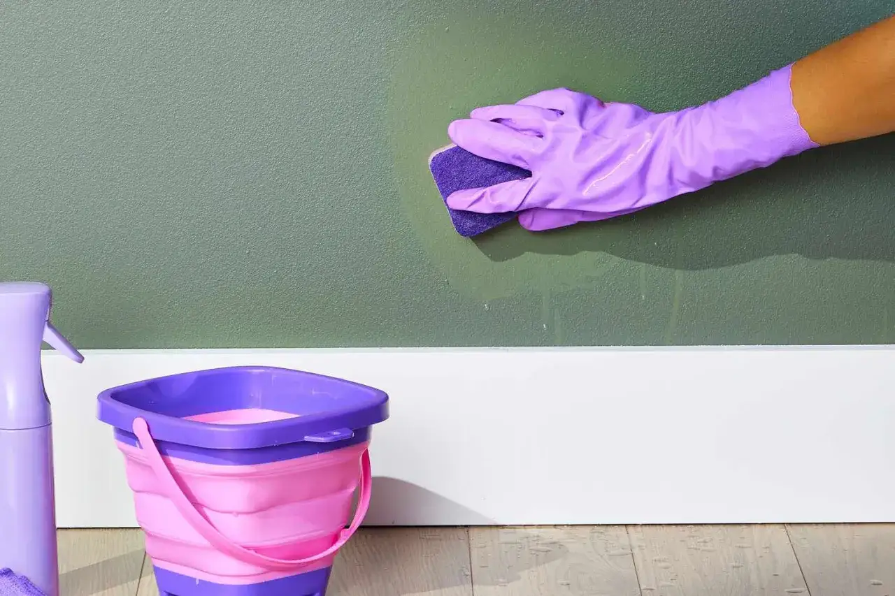 Mit Hausmittel Filzstift entfernen: Liliafarbene Handschuhe und Schwamm reinigen die Wand. Ein faltbarer Eimer und Sprühflasche stehen bereit.
