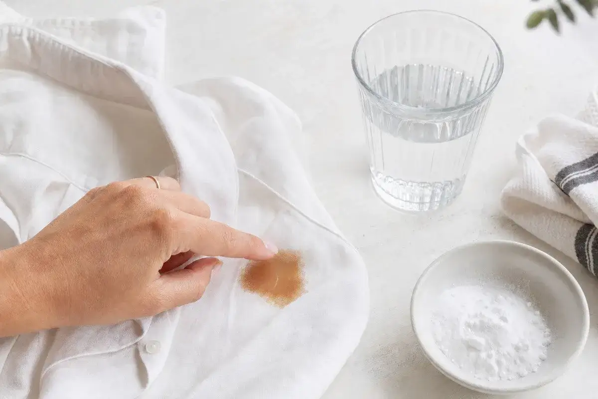 Ein Finger zeigt auf einen braunen Kaffeefleck auf einem wei&szlig;en T-Shirt. Daneben stehen ein Glas Wasser und eine Schale mit Natron.