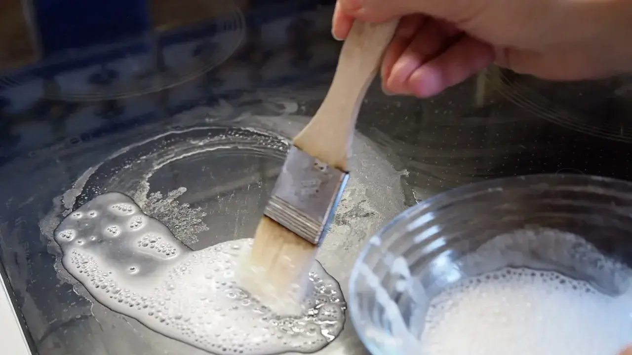 Backpulver als Reinigungsmittel: Eine Hand trägt mit einem Pinsel eine schaumige Paste auf ein Ceranfeld auf, um hartnäckige Flecken zu entfernen.