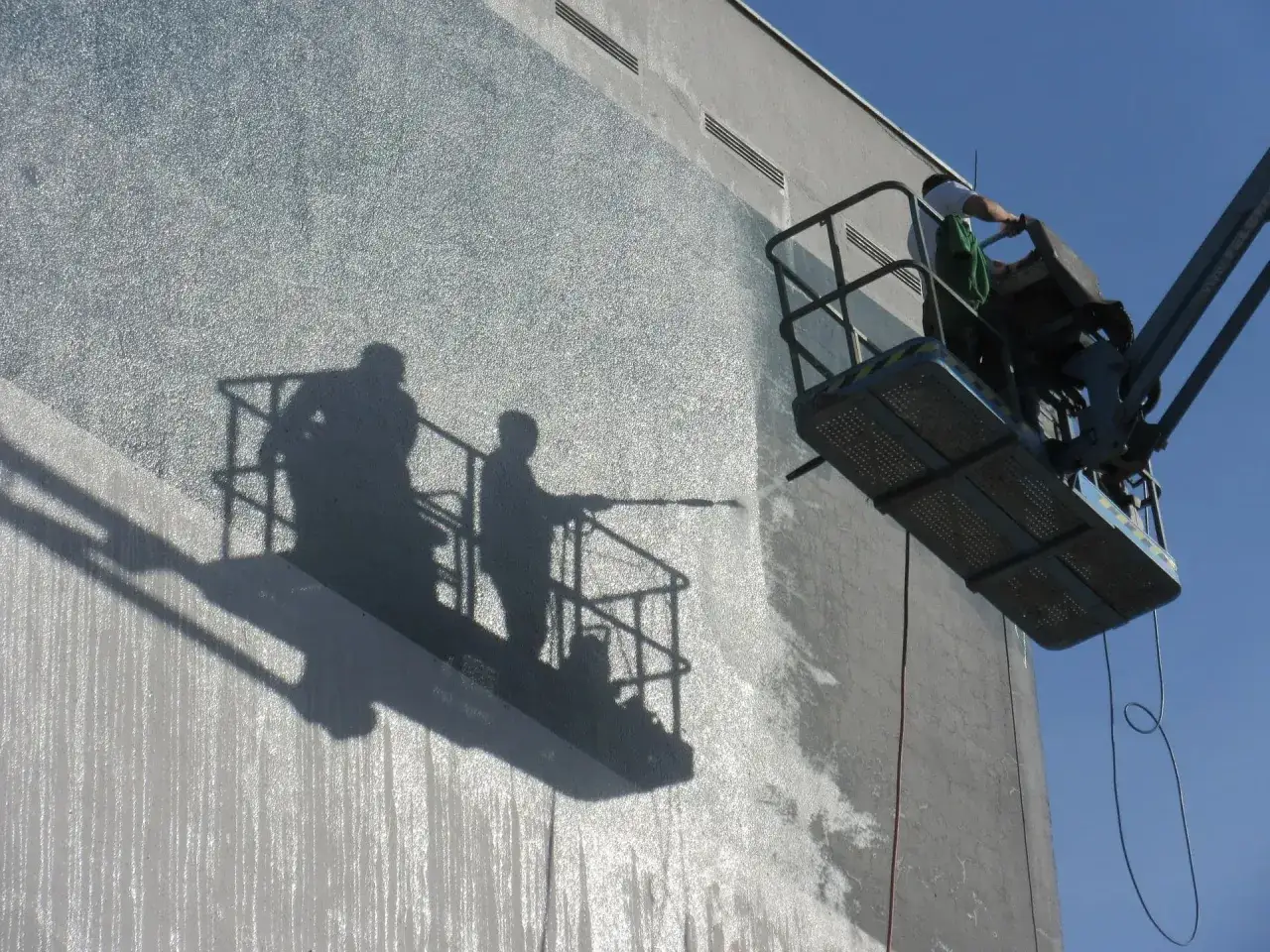 Mann in Hebeb&uuml;hne reinigt mit Hochdruckreiniger die Fassade. Das Selbermachen spart Geld.