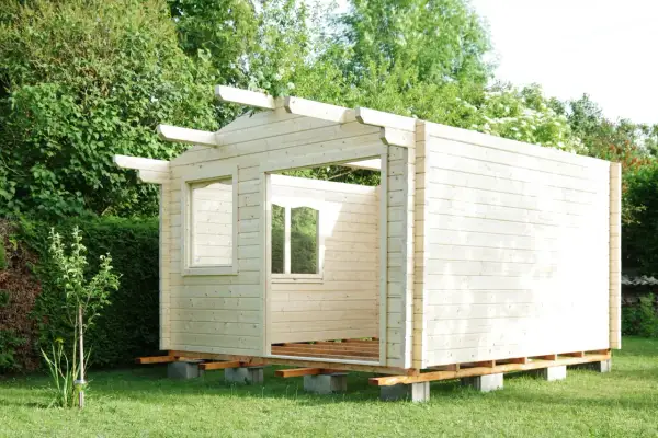Ein Rohbau für ein Gartenhaus, das man selber bauen kann, steht im Grünen. Die Holzwände sind bereits montiert, Fensteröffnungen sind sichtbar.