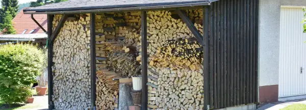 Ein Holzunterstand bauen: Gestapeltes Brennholz wartet unter einem Dach. Daneben eine Garage und grüne Büsche.