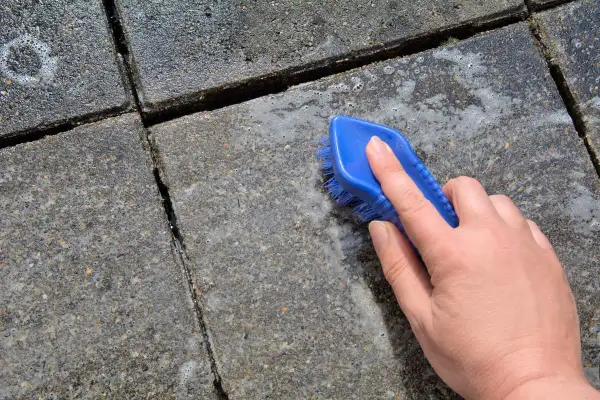 Eine Hand mit blauer Bürste reinigt Betonplatten. Essig greift den Beton an, Schaum entsteht.