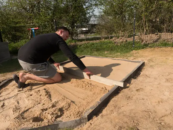 Mann richtet den sandigen Untergrund für den Sandkastenbau mit einer Wasserwaage.
