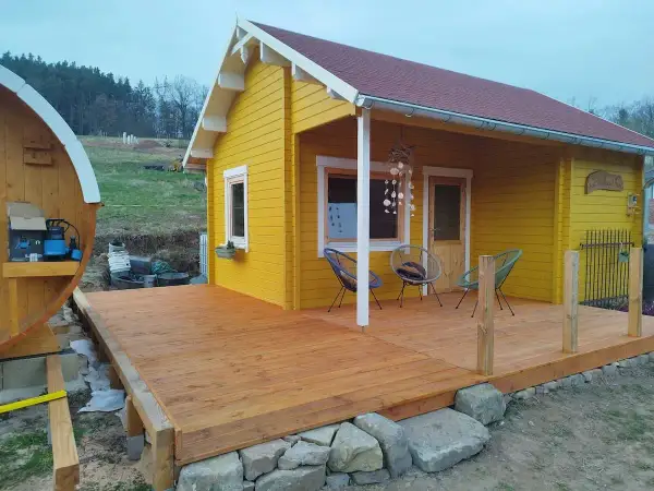 Ein gelber Holzschuppen mit Terrasse, ideal zum schuppen selber bauen. Daneben eine Holzfasssauna.